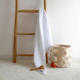 White 4-ply muslin baby blanket draped over wooden ladder beside nursery basket